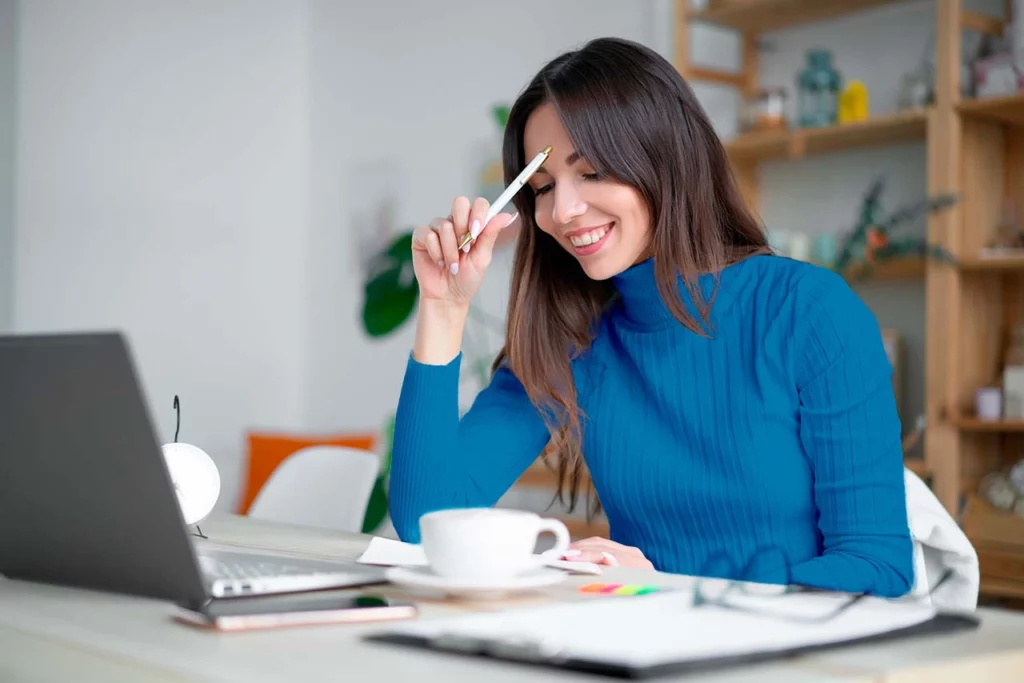 Mujer sonriente con bolígrafo en mano sentada viendo la laptop 