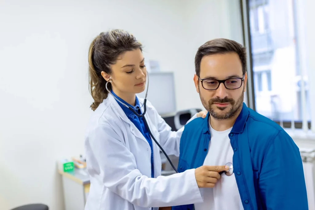 Paciente hombre, recibiendo consulta de medicina general por parte de una doctora que escucha sus pulmones.