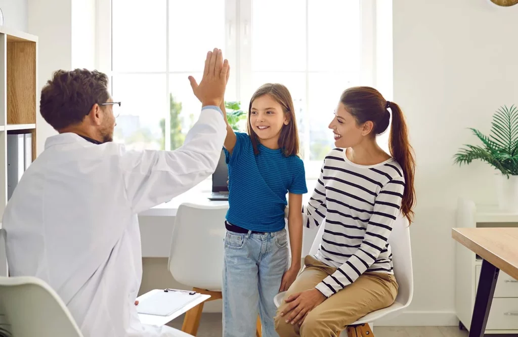 Médico reumatólogo pediatra chocando la mano con su paciente mientras su mamá sonríe.