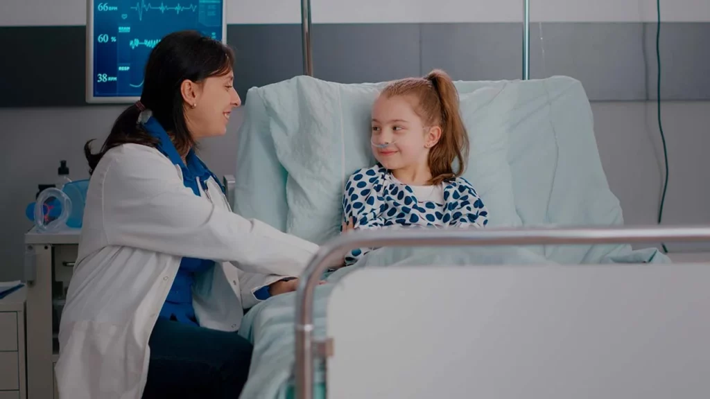 Niña sentada en una cama de hospital, después de su procedimiento de cirugía pediátrica, hablando con su doctora.