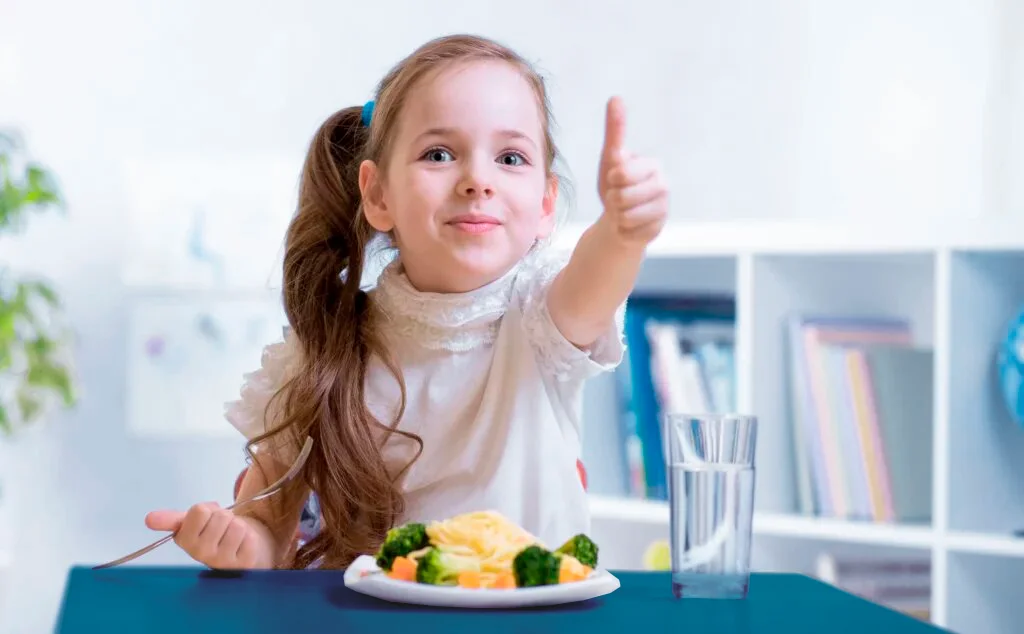 Niña disfrutando del plato del buen comer para niños mientras está sentada a la mesa y levanta el pulgar.