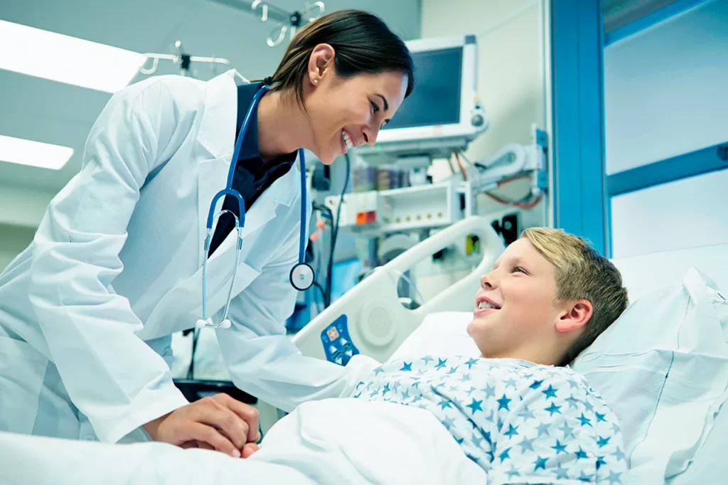 Paciente de cuidados críticos pediátricos acostado en una cama de hospital sonriendo, mientras su doctora lo revisa.