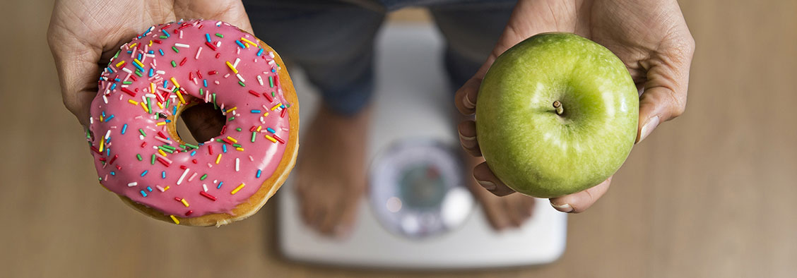 Persona sobre una balanza sosteniendo una rosquilla en una mano y una manzana verde en la otra
