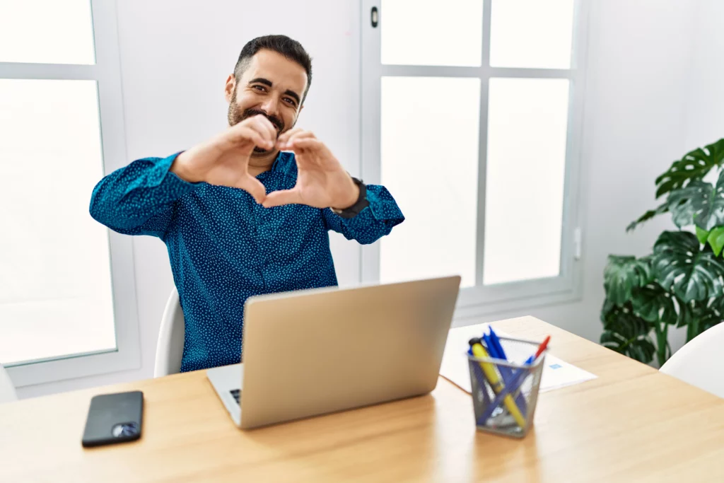 Joven trabajando en oficina con laptop sonriendo haciendo un símbolo de corazón