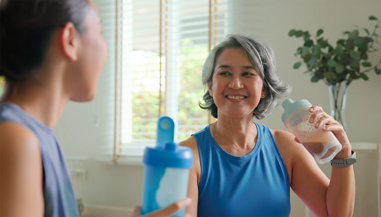 Mujer de mediana edad tomando un batido de proteína, mientras le sonríe a su hija. Mujer de mediana edad tomando un batido de proteína, mientras le sonríe a su hija.