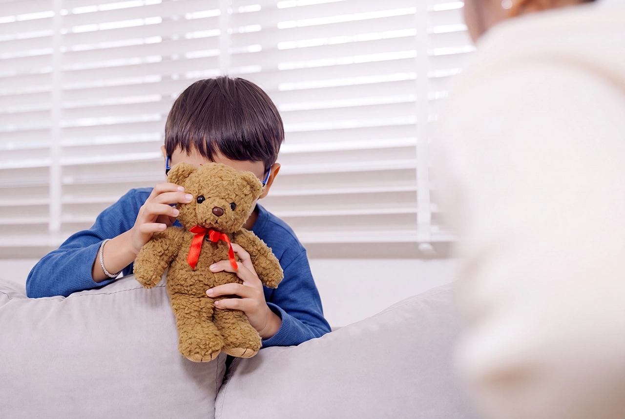 Niño cubriendo su rostro con un oso de peluche en consulta, como parte de sus síntomas de TDAH. Niño cubriendo su rostro con un oso de peluche en consulta, como parte de sus síntomas de TDAH.