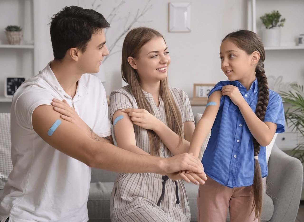 Familias de papá, mamá e hija, con una tira adhesiva en el brazo para indicar que recibieron nuevas vacunas como parte de su plan de salud.
