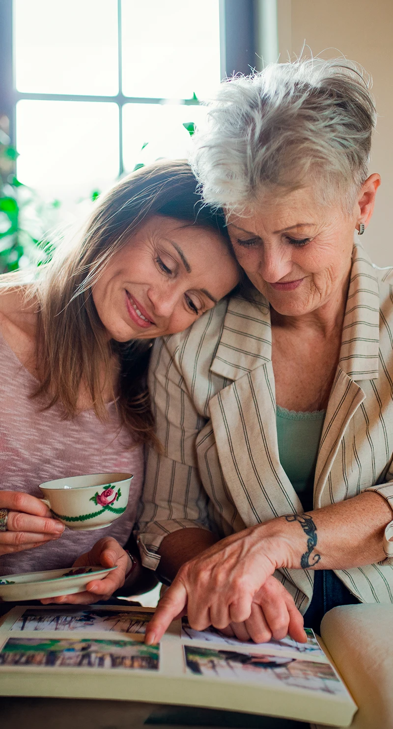 Madre e hija sonrientes en la cocina y viendo fotos, representando el factor de riesgo de antecedentes familiares de cáncer de mama. Madre e hija sonrientes en la cocina y viendo fotos, representando el factor de riesgo de antecedentes familiares de cáncer de mama.